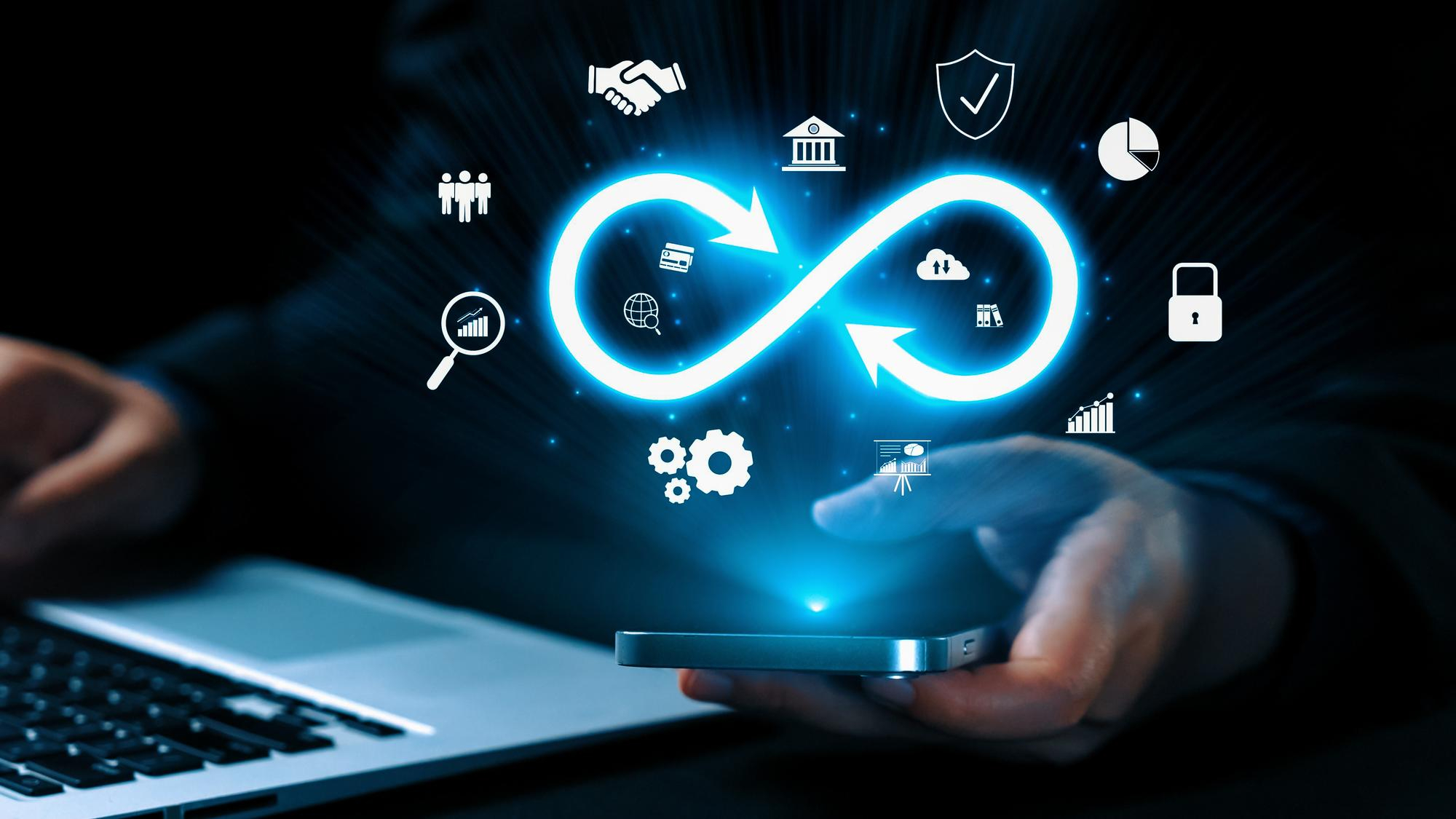 Hand holding a smartphone with a glowing DevOps infinity loop and tech icons above, in front of a laptop.