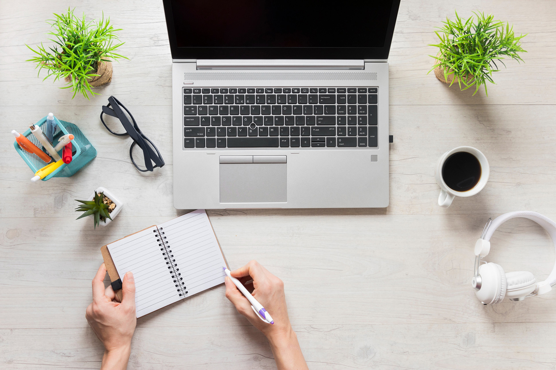 A person writes in a notebook at a desk with a laptop, coffee, headphones, pens, glasses, and potted plants. No faces are visible