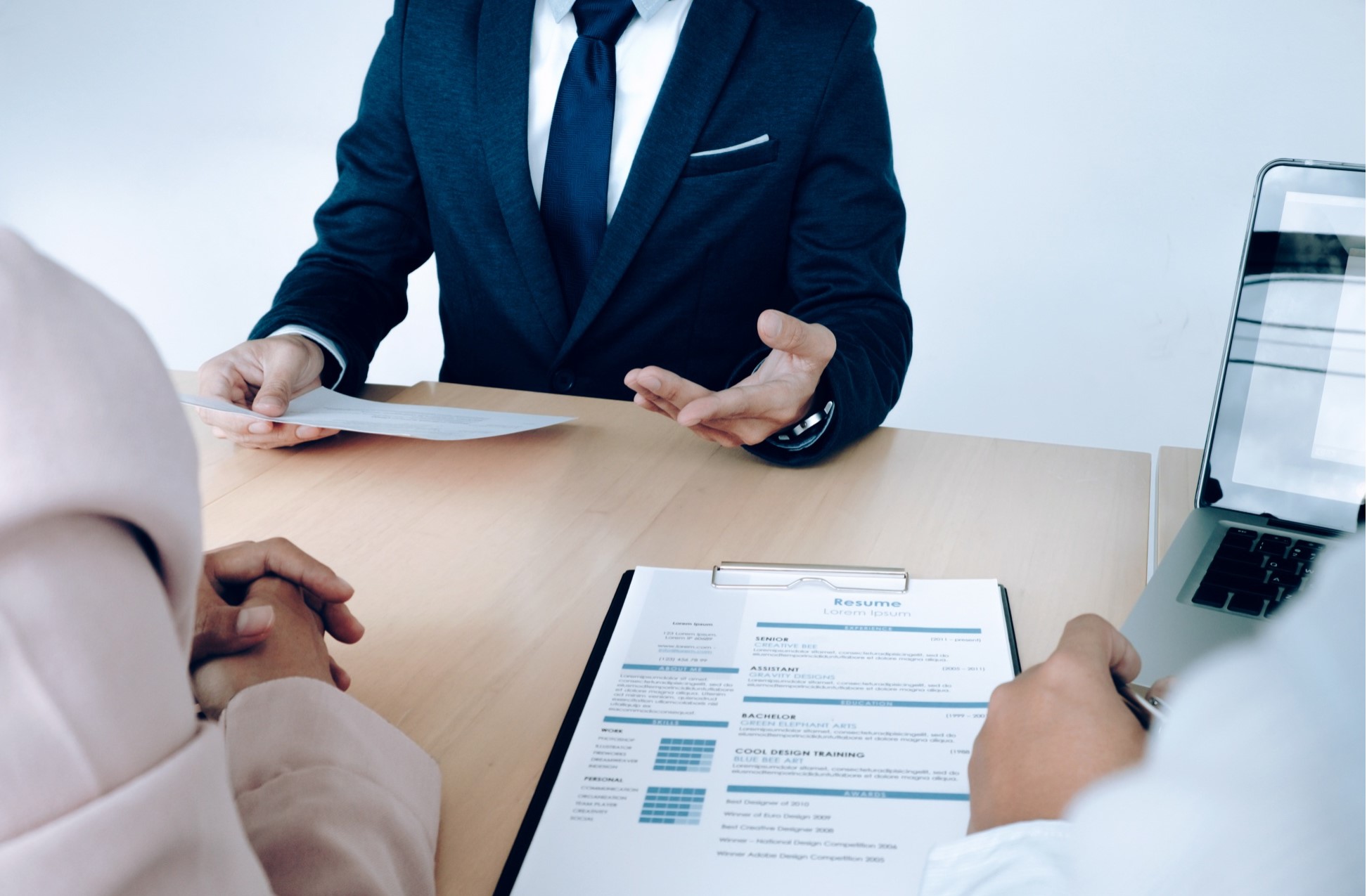 Businessperson in a suit interviewing a candidate across a desk, with a resume and laptop visible.