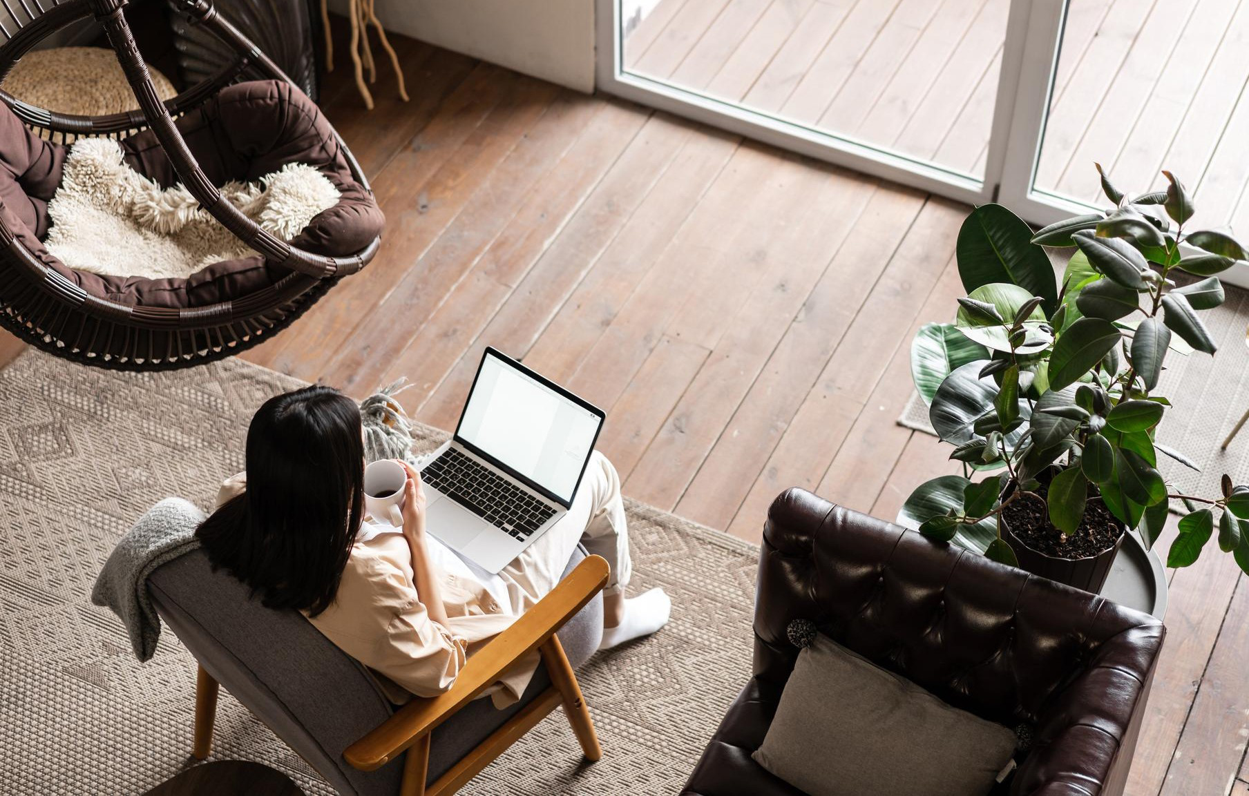 A person sits in an armchair with a laptop and mug in a cozy living room; no faces are visible.