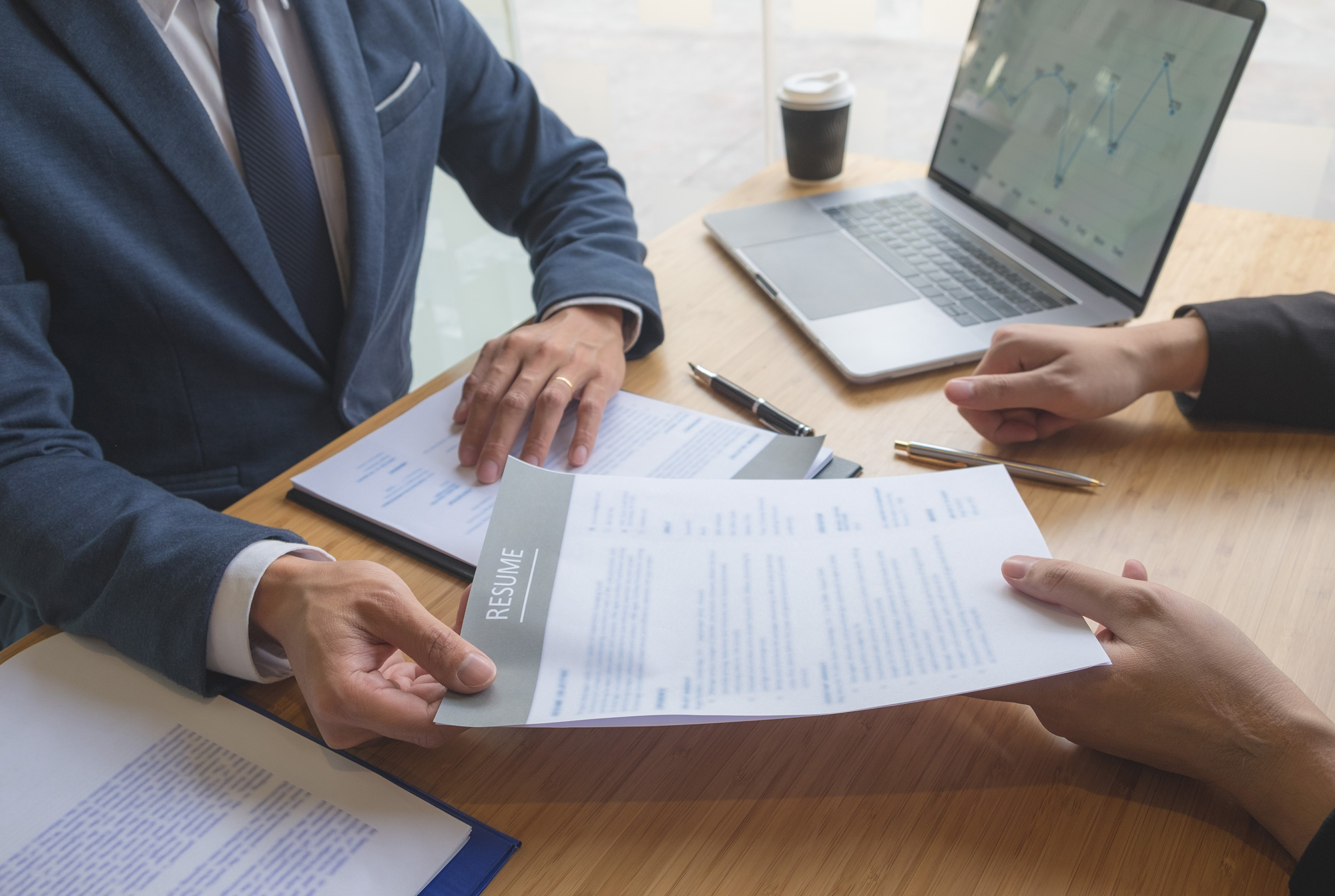 Hands of interviewers and a candidate exchanging a resume across a desk, laptop and documents in view, faces out of frame.